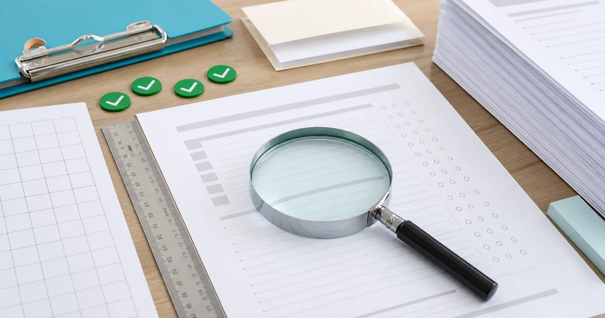 A worksheet quality-control scene with a magnifying glass, spacing ruler, green checks, answer envelope, and page stack.